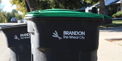 Trash bins with green lids labeled "Brandon The Wheat City" on a sunny street.