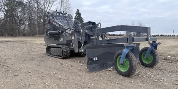 Compact tracked vehicle with blade and wheels on a dirt path in a rural area.