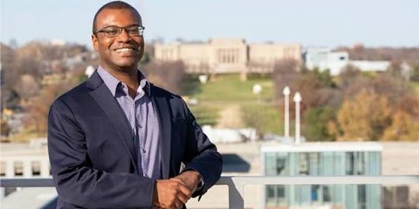 Man smiling outdoors with buildings in the background.
