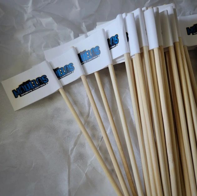 Wooden sticks with white flags featuring the Modelo Sports Cantina logo, neatly arranged 
