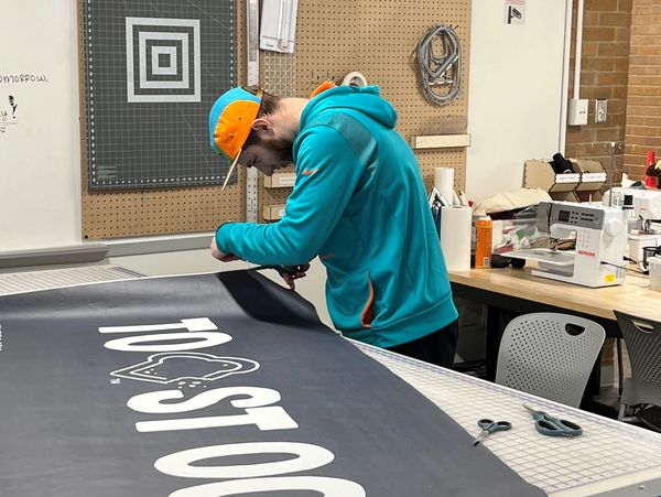 Man in turquoise hoodie cutting a large printed banner in a workshop.