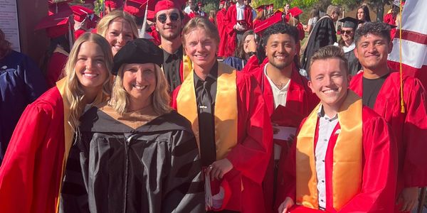 Group of happy graduates and a faculty member celebrating outdoors in red gowns and caps.