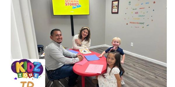 Two adults and two children smiling around a red table in a kids' learning room.