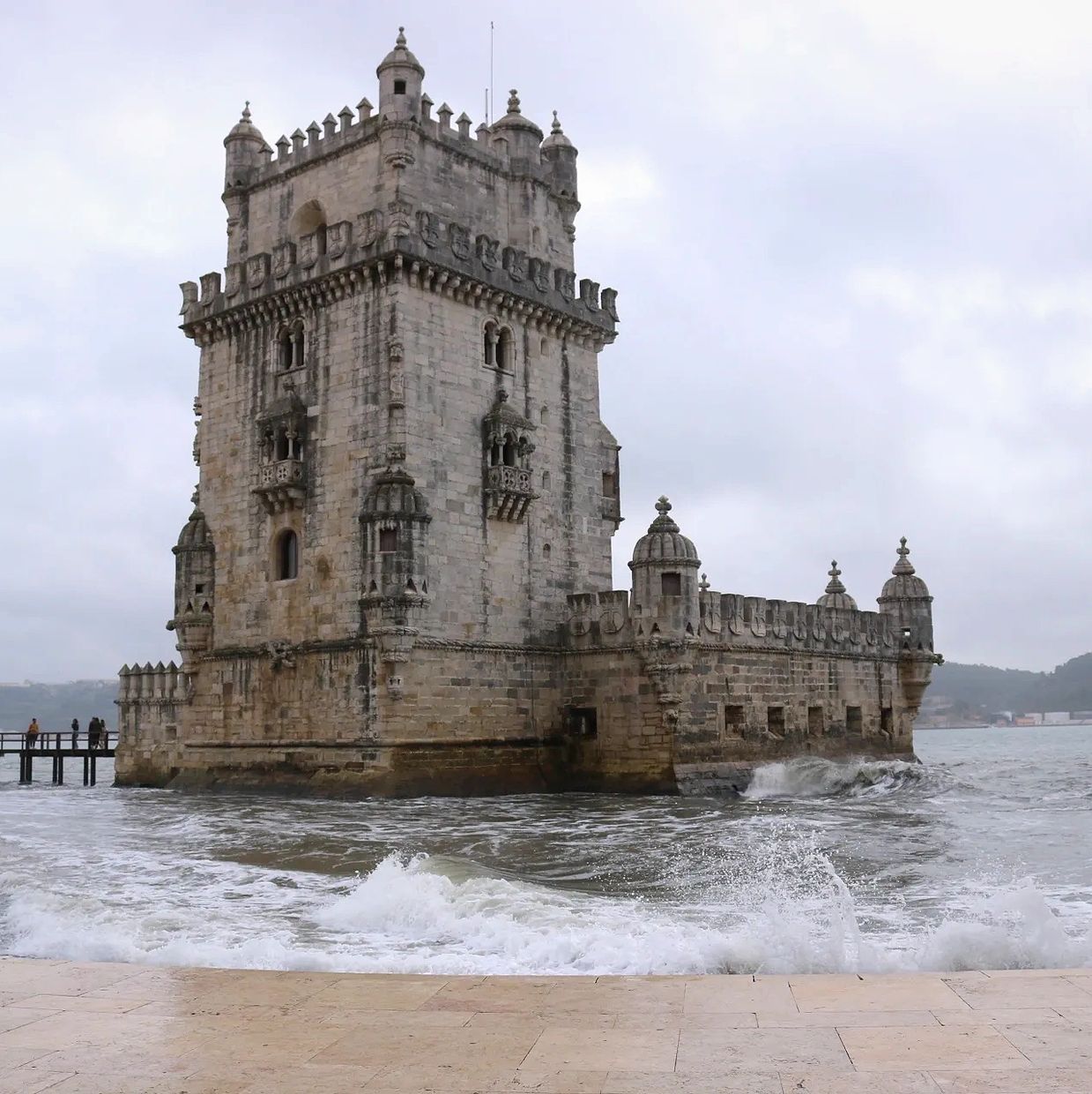Picture of Belém Tower on a cloudy day with waves crashing against marble walkway