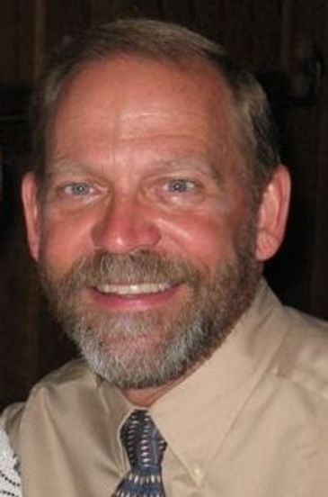 Smiling man with a beard wearing a beige shirt and patterned tie.