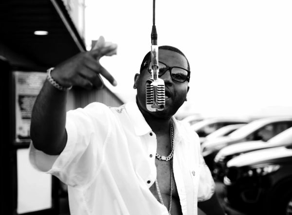 Man in glasses and chain gesturing near a hanging microphone in black and white.