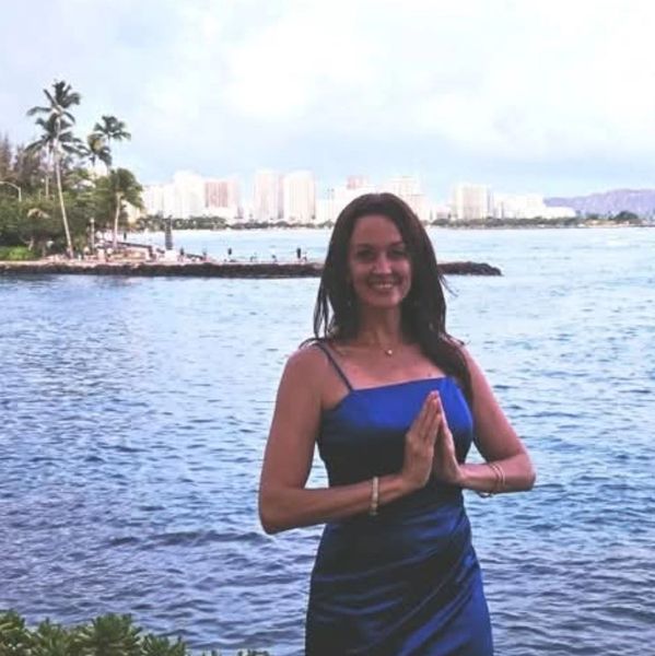 Woman in a blue dress posing by the ocean with hands in a prayer gesture.