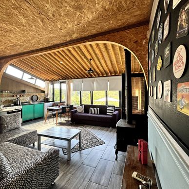 Cozy modern living room with patterned chairs, a purple couch, and a wood stove under a wooden ceiling.