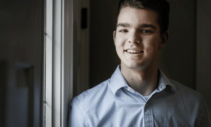 Young man smiling beside a window in a blue shirt.