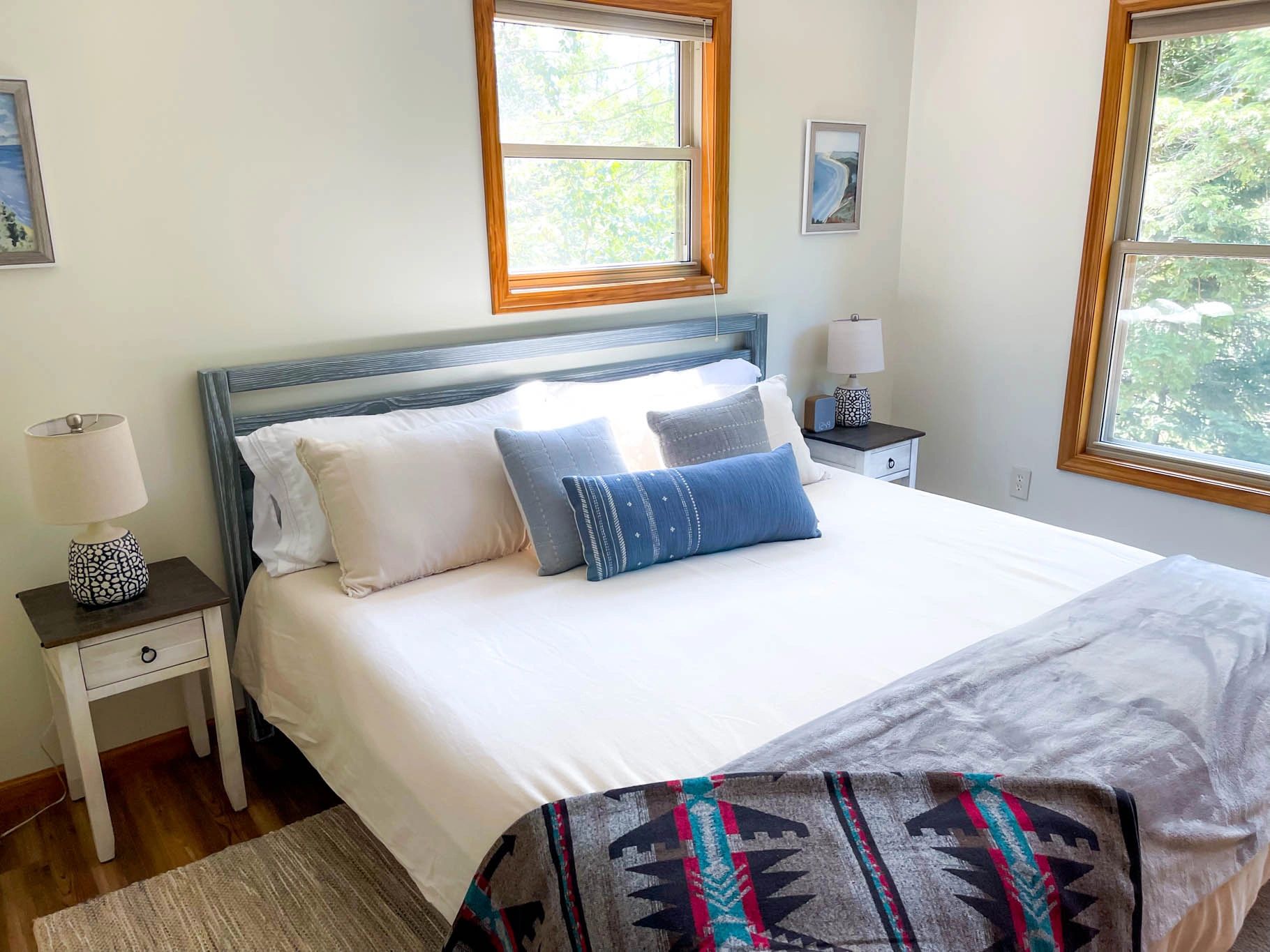 A neatly made bed in a room with white walls, two windows, and a colorful blanket at the foot.