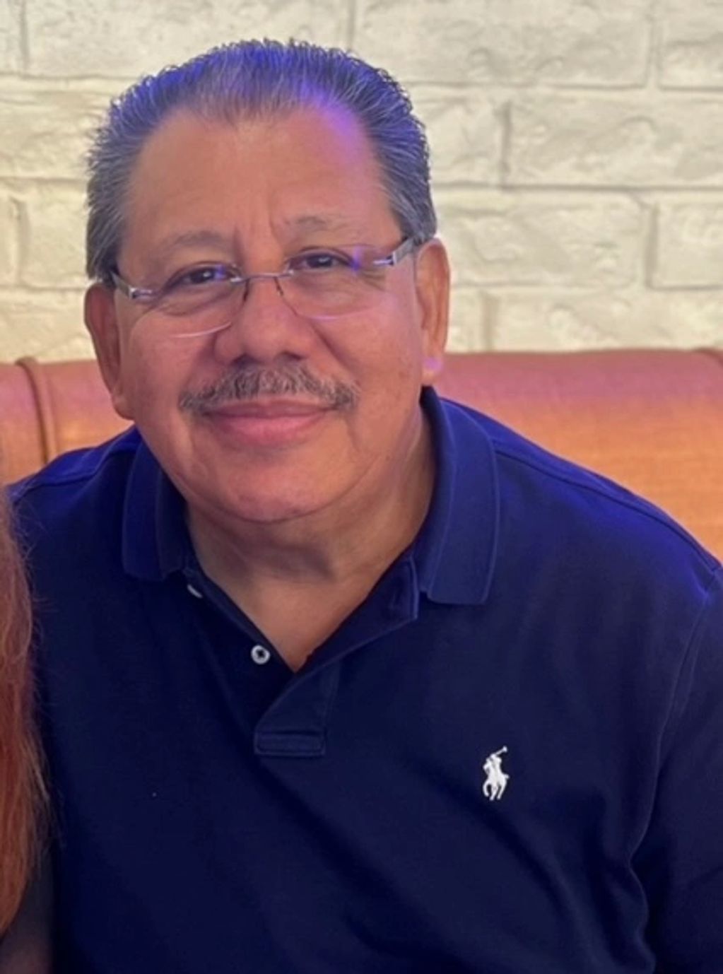 Smiling man wearing glasses and a navy blue polo shirt sitting indoors.
