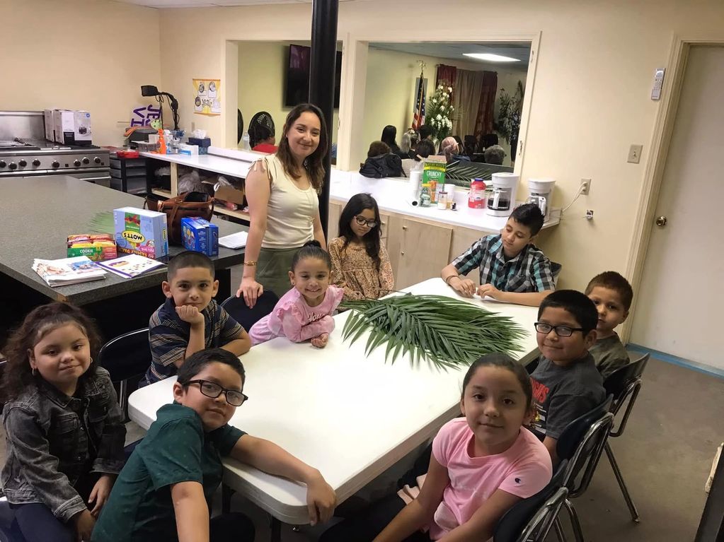 A group of children and a woman around a table with palm leaves indoors.