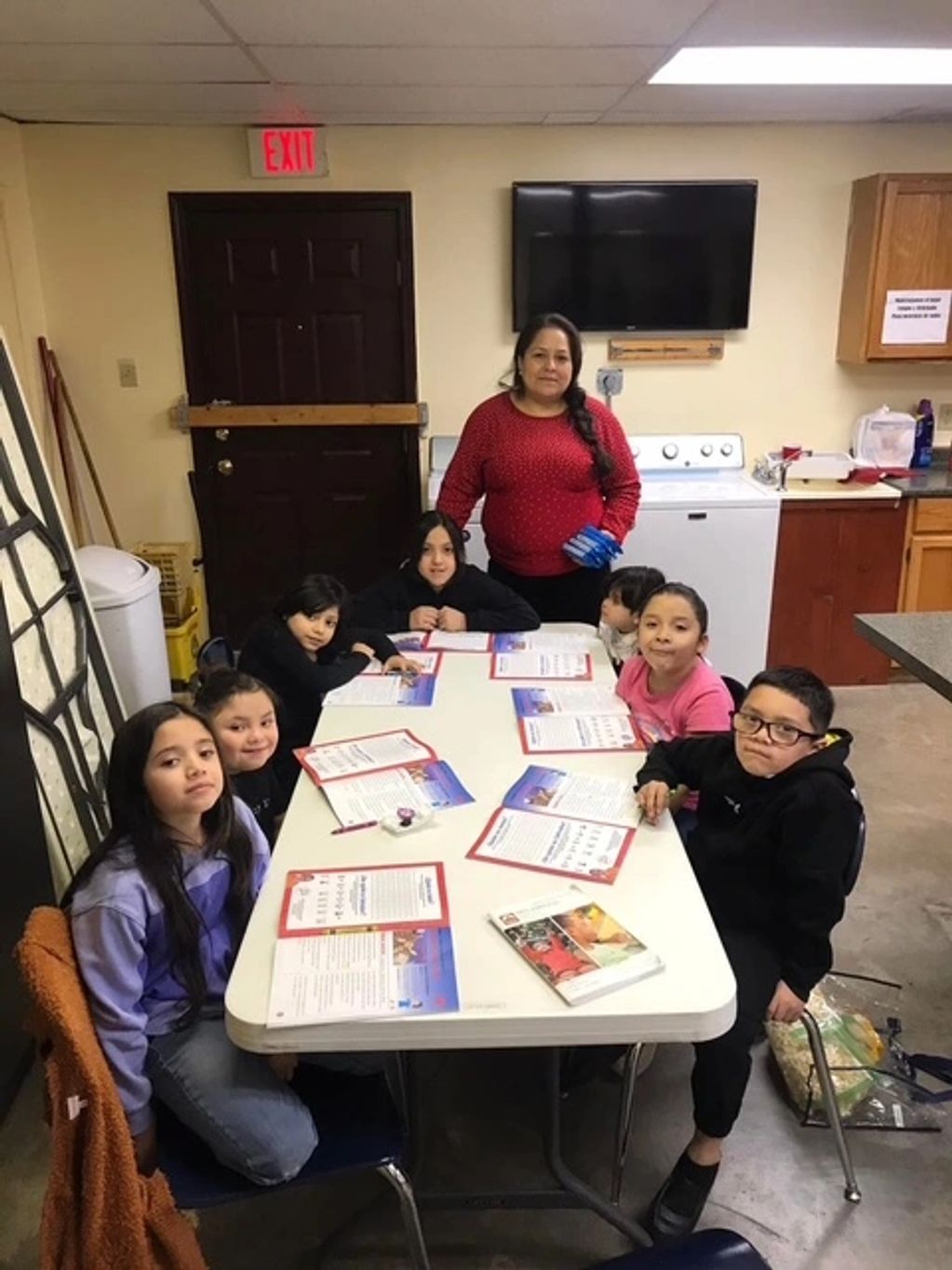 A group of children and a woman around a table with papers in a classroom-like setting.