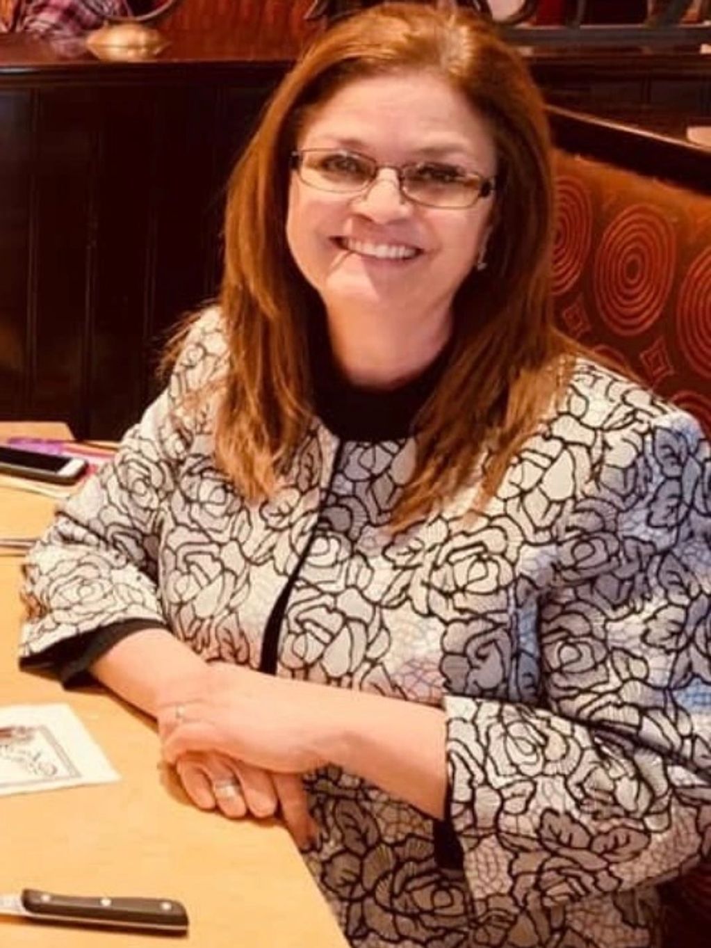 Smiling woman with glasses sitting at a table in a patterned jacket.