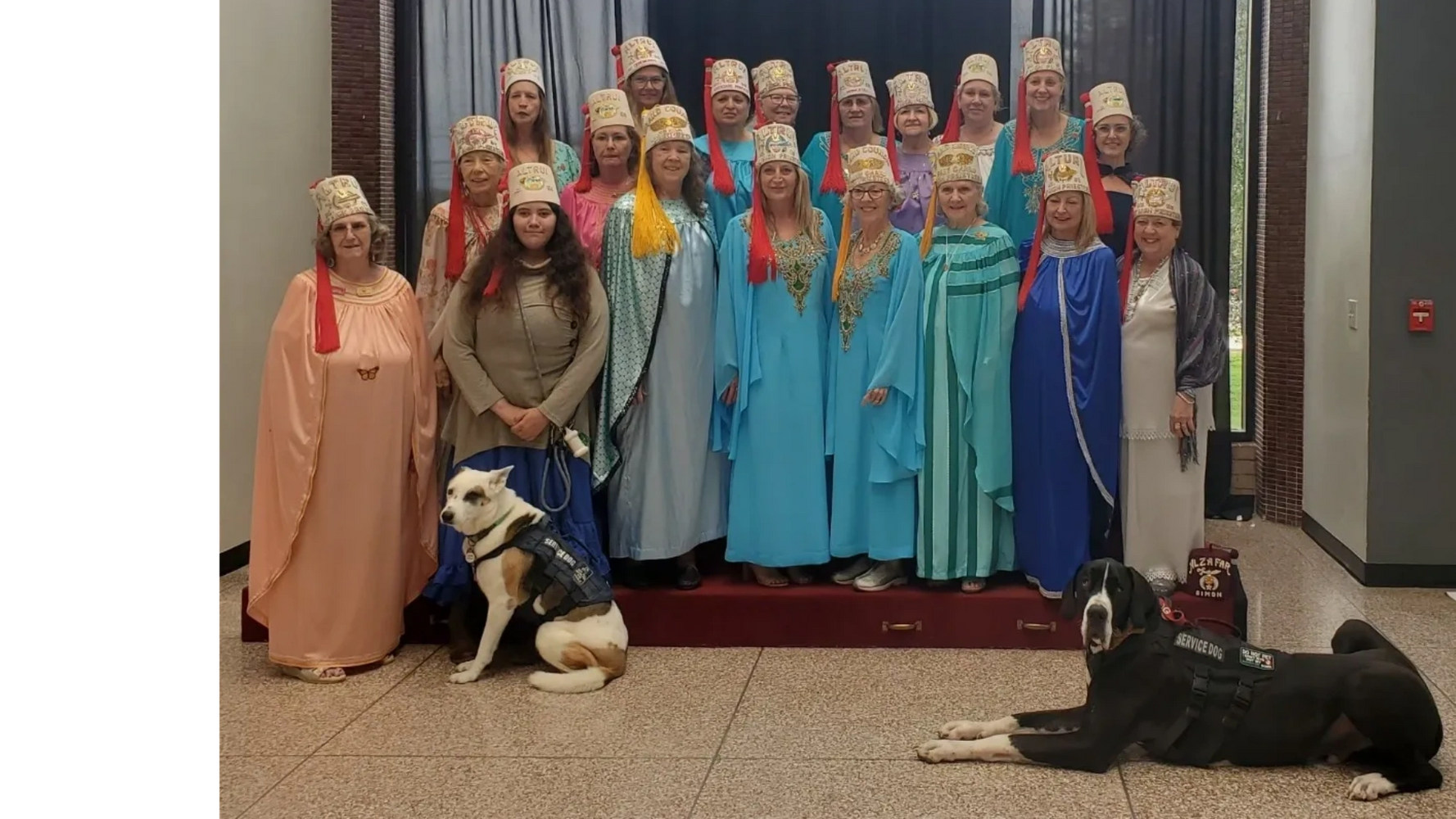 Group of women in ceremonial robes and hats posing with two service dogs indoors.