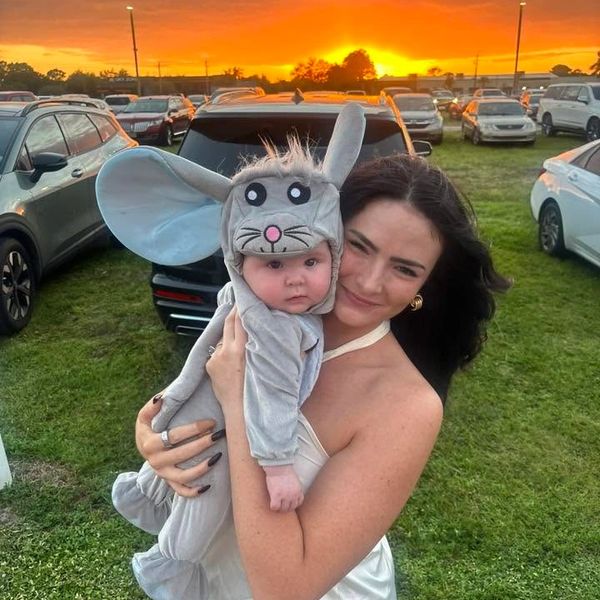 Woman holding baby dressed in a cute mouse costume at sunset in a parking lot.