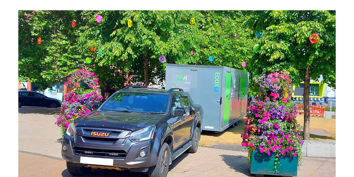 Black Isuzu pickup truck parked near flower displays and a trailer in a sunny outdoor setting.