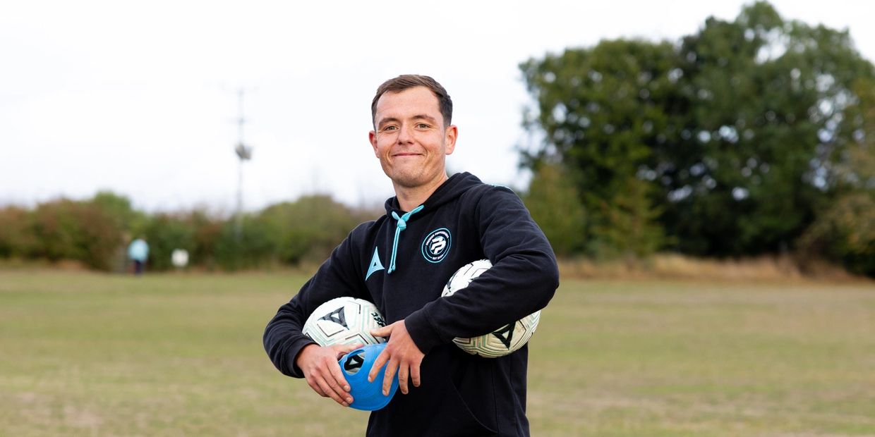 Man holding three sports balls and smiling outdoors.
