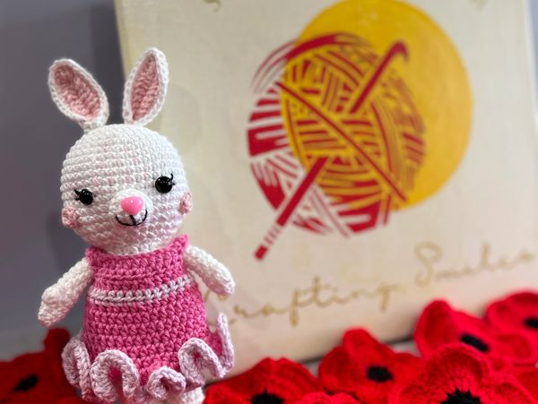 Crocheted bunny in a pink dress surrounded by red crocheted flowers.