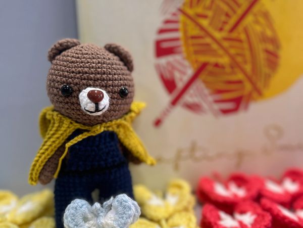 Crocheted bear with yellow cape standing among colorful crocheted flowers.