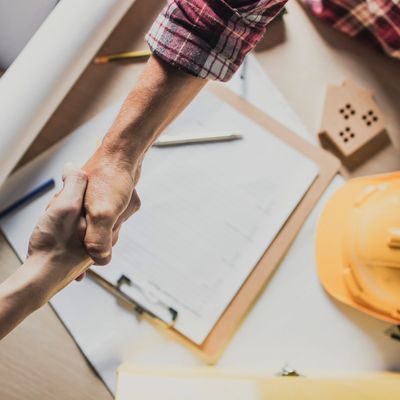 Two people shaking hands over architectural plans and a safety helmet.
