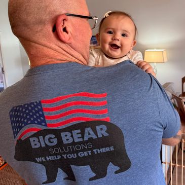 A man holds a happy baby, wearing a Big Bear Solutions shirt with an American flag.