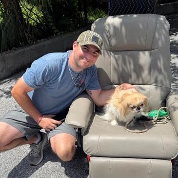 Man kneeling beside a dog sitting on a recliner outdoors on a sunny day.
