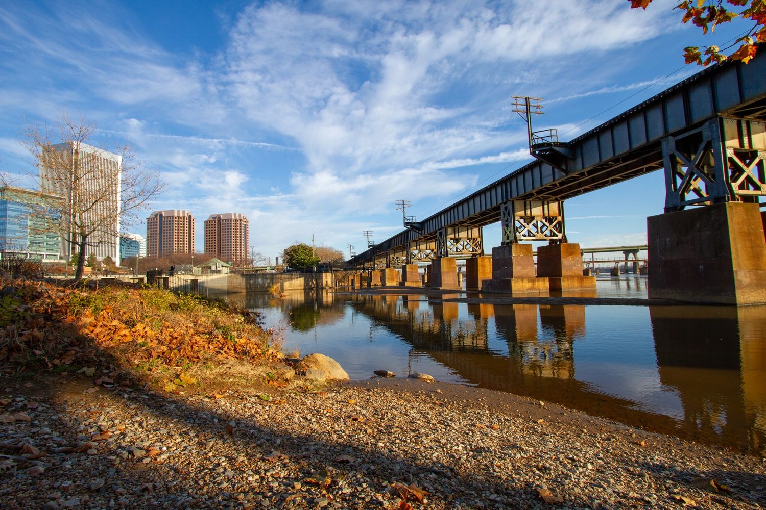 Railway bridge over calm river with city buildings in the background on a sunny day.