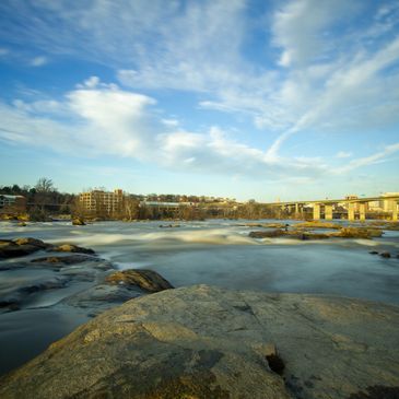 Calm river with rocks, buildings, and a bridge under a partly cloudy sky.