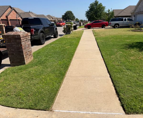 A suburban sidewalk flanked by green lawns and parked vehicles.