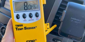 Hand holding a yellow digital temperature meter inside a vehicle.