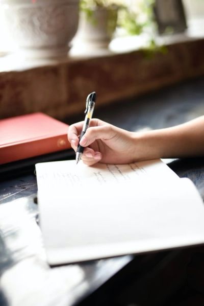 Person writing with a pen in a notebook by a window.