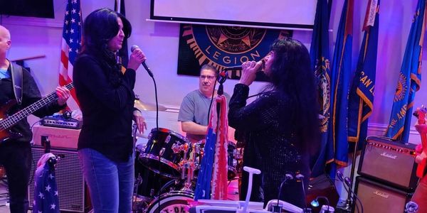 Two women singing on stage with a drummer and bassist in the background.