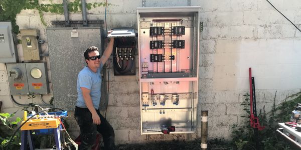 Man working on an electrical panel outdoors against a white brick wall.