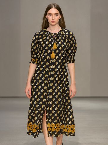 Model wears a black midi dress with yellow floral patterns and tassels.
