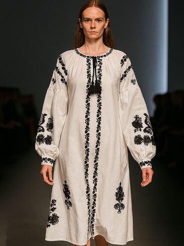 Model wearing a white dress with black floral embroidery and long sleeves on runway.