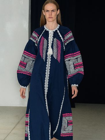 Model wearing a navy blue dress with white and pink embroidery walking on runway.