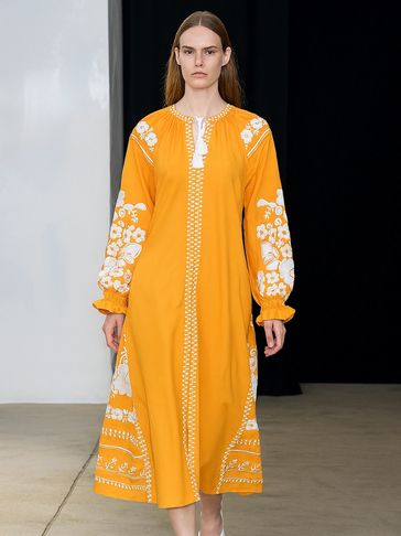 Model walks runway in a vibrant yellow dress with white floral embroidery and white shoes.