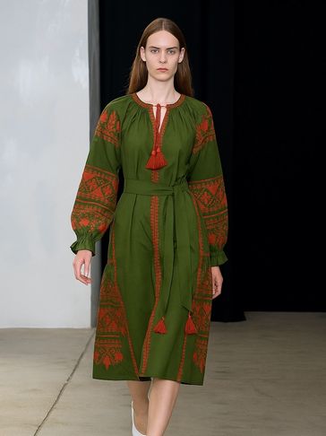 Model walking in a green dress with red embroidery and white shoes.
