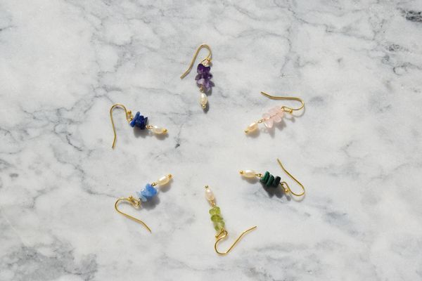 Six pairs of colorful gemstone and pearl earrings arranged in a circle on marble.