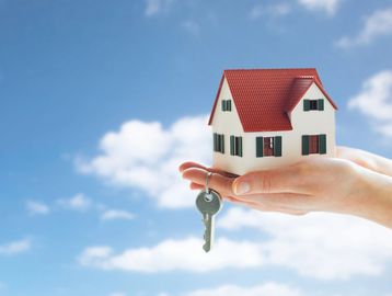 Hand holding a small house model with a key against a blue sky.