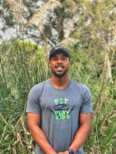 Man wearing a Nike cap and 'Fit 2 Play Kids' shirt outdoors.