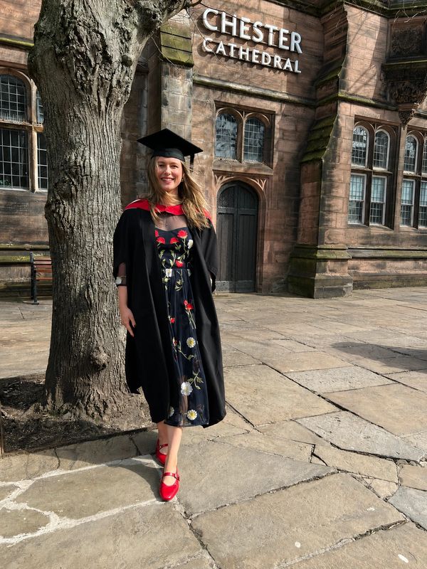 A smiling graduate in a black gown and cap stands outside Chester Cathedral.