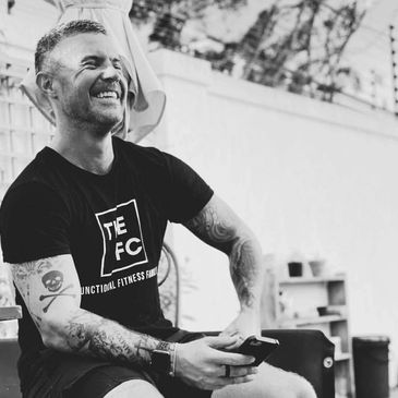 Man with tattoos smiling while holding a phone, sitting outdoors.