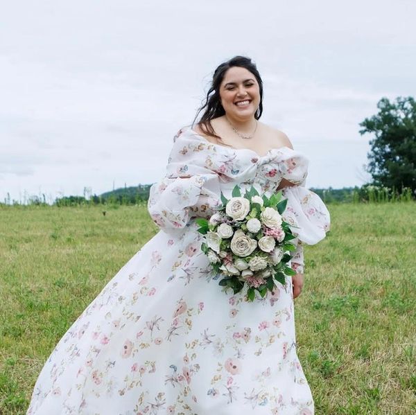 Smiling bride in flowered wedding dress standing in a field with a white and pink wood flower bouque