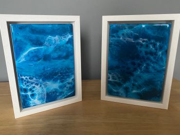 Two framed blue abstract fluid art pieces on a wooden surface.