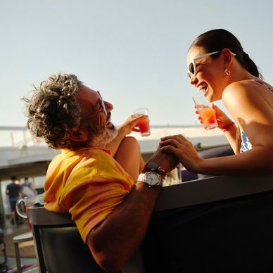 Man and woman sitting together on a Virgin Voyages ship laughing and holding hands with a drink