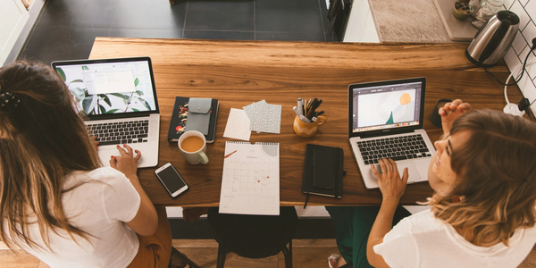 Two women working on laptops at a wooden table with coffee and stationery.