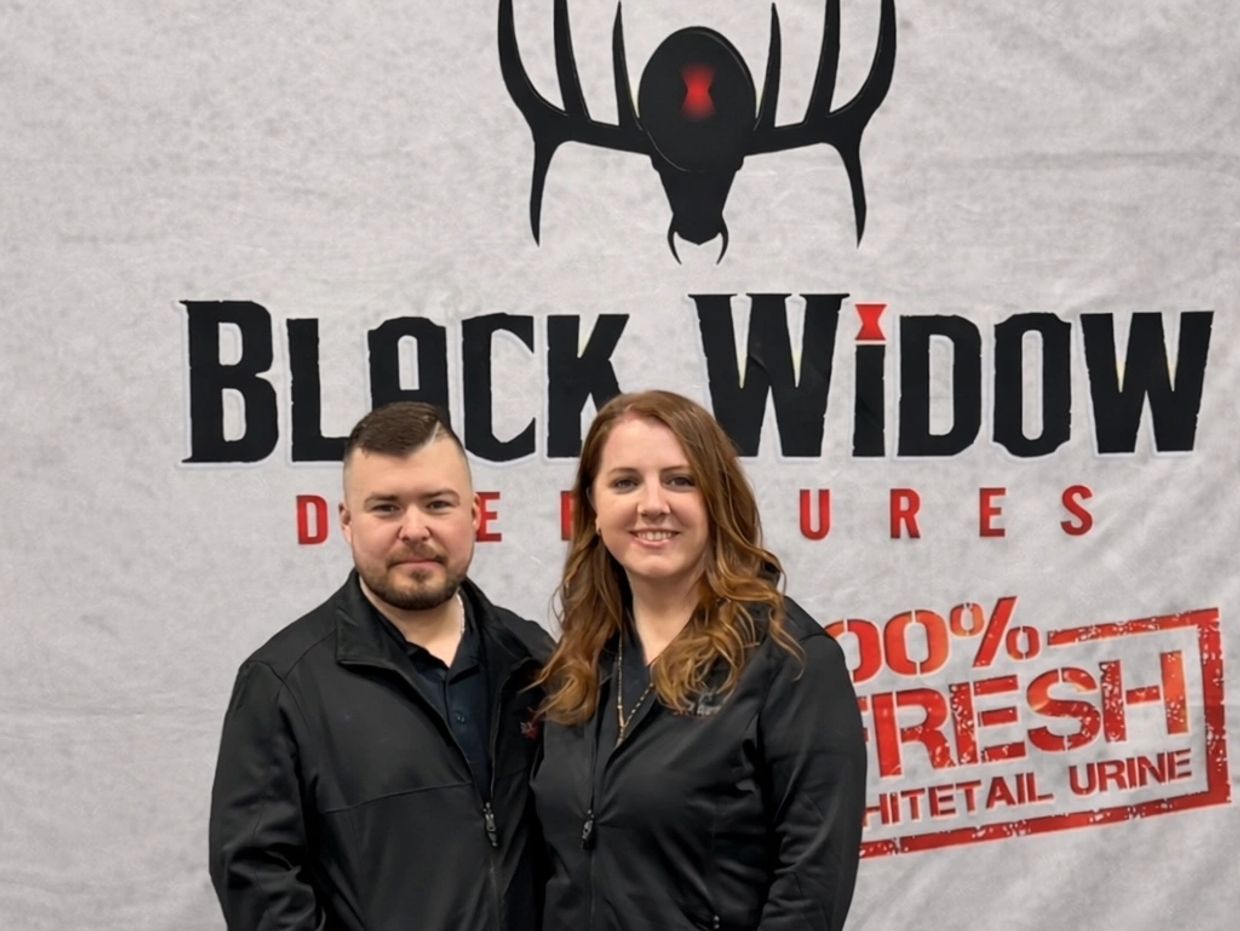 Two people posing in front of a Black Widow Deer Lures banner.