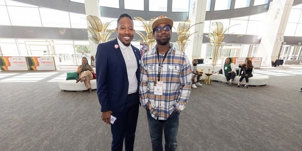 Two men posing at a spacious indoor event with people seated in the background.